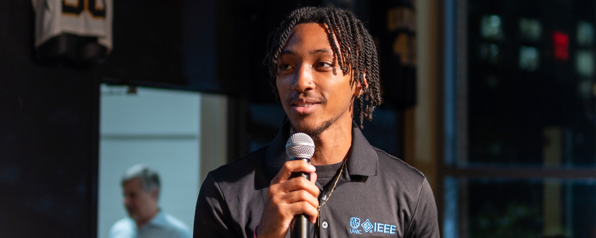 A young man stands in a black polo shirt talking into a microphone