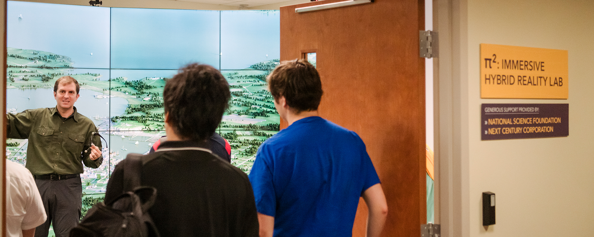 man standing in front of large screen as several students stand in doorway