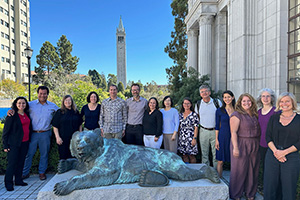 A large group of people gather outside behind a metal statue of a bear. A tall tower is in the distance.