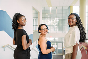 Three women stand at top of stairs looking at camera with window view to outside in the distance.