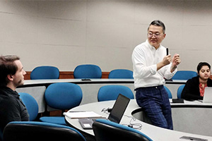 Man in glasses talks with students seated in circular rows of desks and chairs in lecture hall.