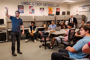 A group of college students sitting around rectangular desks. There is a person standing near the left hand side who is pointing up towards something out of camera.