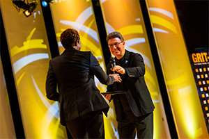 Two people on a stage, one of them has their back turned away from the camera. The man on the right is extending a microphone to woman on the left.