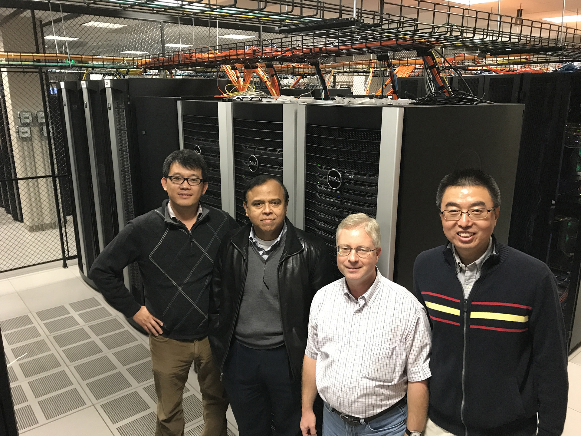 Four men facing camera standing in foreground with high performance computers in background.