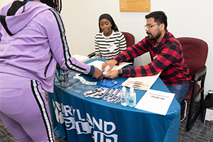 two Pre-law student volunteers sit at a table with a blue and white table cloth with the letters Maryland Legal Aid in white