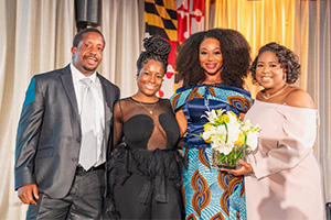 Four people smile on stage and look at camera with Maryland flag in background
