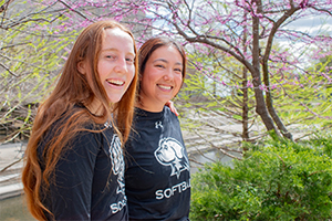 Two girls, one with red hair and one brunette, smiling in front of a flowering tree with arms around each other. Both are wearing black UMBC Athletics shirts.