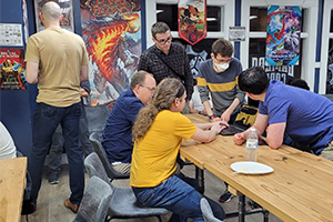 People gather around a table with a laptop in a game store, surrounded by gaming posters.