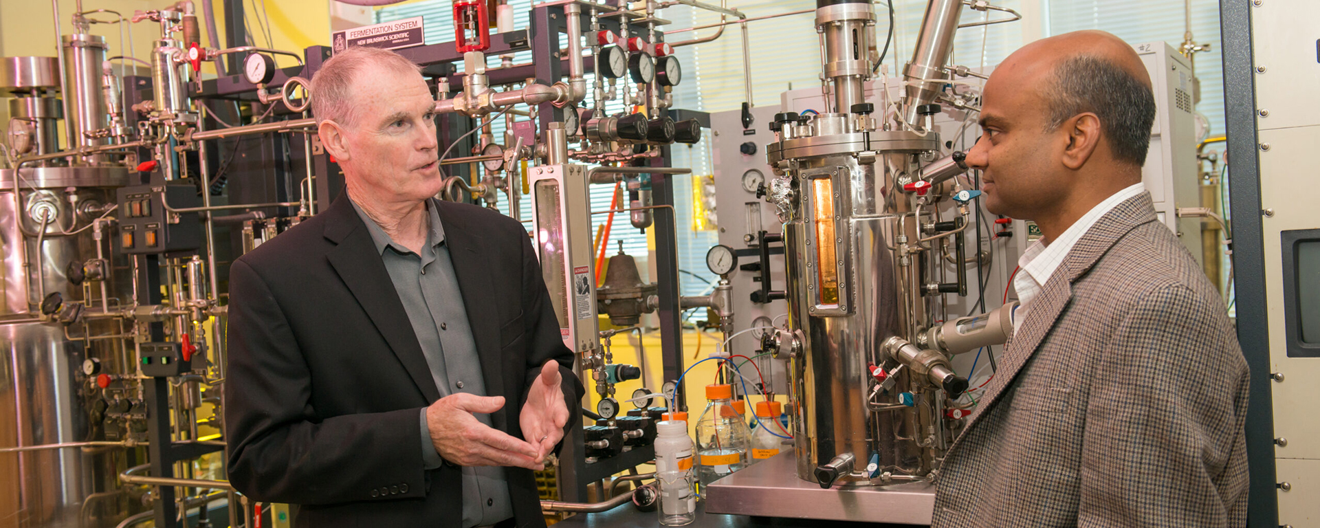 two men in conversation in front of a complicated set of tubes, flasks, and sensors