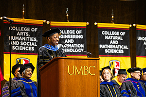 woman stands podium with letters U-M-B-C on front and seated audience members looking out toward camera