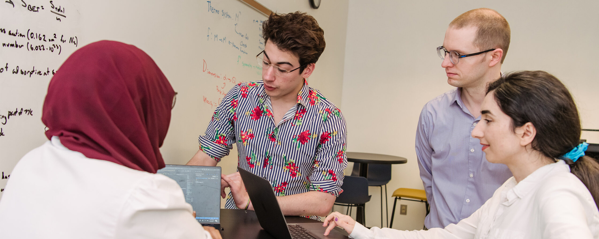 Group in discussion gathers around computers. Equations on whiteboard in background.