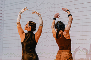 Two dancers perform in front of screen displaying wavy line signals.