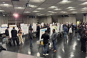 People mingle in a large room around posters mounted on tables.