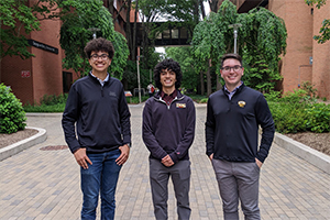Three students wearing UMBC shirts stand outside, with academic buildings in background.