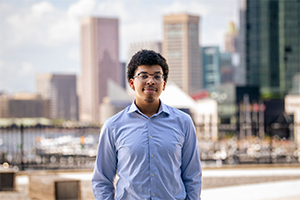 Student in button-down shirt stands in front of skyscrapers, waterfront, and boats.