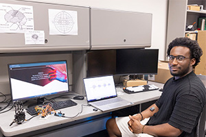 A man sits next to table with computers, electronic wiring and a robotic hand.