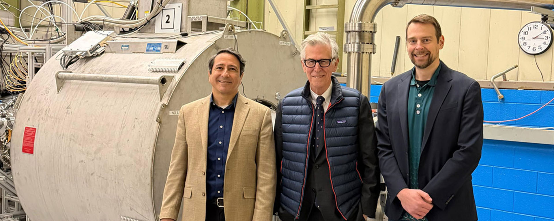 Three men stand in an open laboratory space in front of a large metal machines connected to wires and tubes.
